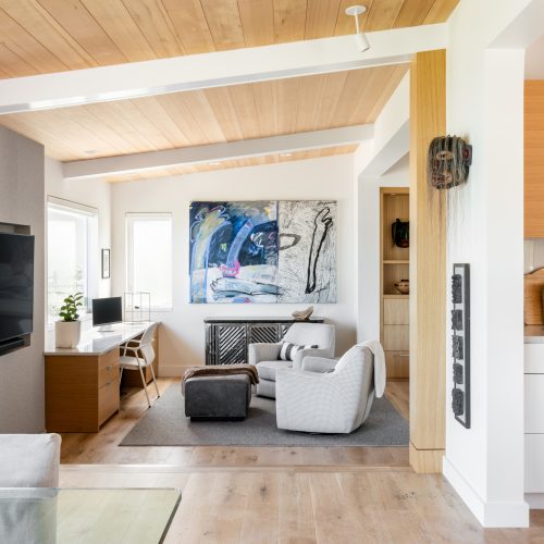 Modern living room with light wood flooring and ceiling, featuring a TV on the left wall. There's a desk with a chair, and two armchairs with an ottoman on a gray rug. Abstract art hangs on the back wall. A partial view of a kitchen is on the right.