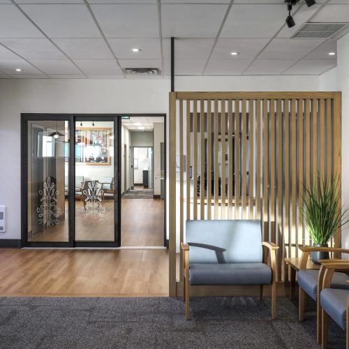 A modern waiting area with wooden flooring and carpet, featuring two blue armchairs, a wooden slat partition, a potted plant, and vibrant artwork on the wall. Glass doors lead to another room, and overhead lighting illuminates the space.