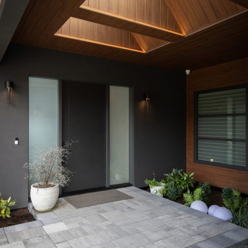 Modern front entryway with potted plants, a large dark door, frosted glass panels, gray stone tiles, and a wood-paneled ceiling featuring square skylights and recessed lighting.
