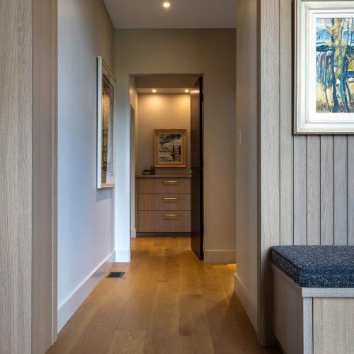 A hallway with light wood flooring and walls, leading to a small, lit room with cabinets. Framed artwork hangs on the walls, and there is a built-in bench with a dark cushion on the right.