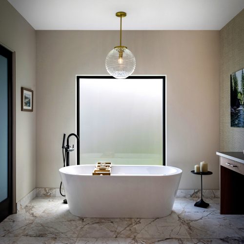 Modern bathroom with a freestanding white bathtub centered below a large frosted window, marble tile floor, a black floor-mounted tub filler, a round side table with candles, and a glass pendant light overhead.