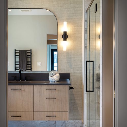 Modern bathroom with light wood vanity, black fixtures, a large mirror, wall sconce, towel radiator, marble floor, and a glass shower enclosure. Neutral color palette with contemporary design elements.