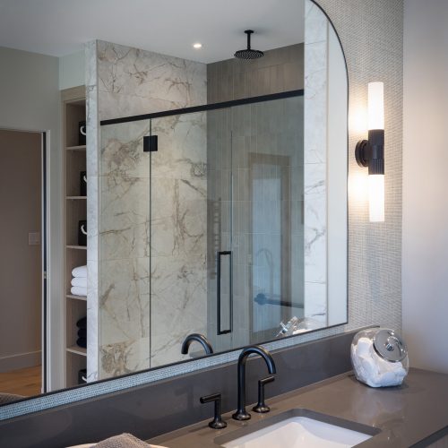 A modern bathroom with a large mirror above a gray countertop, black faucet, round tray with folded towels, wall sconce, and a glass-enclosed shower with marble walls and a black rainfall showerhead.