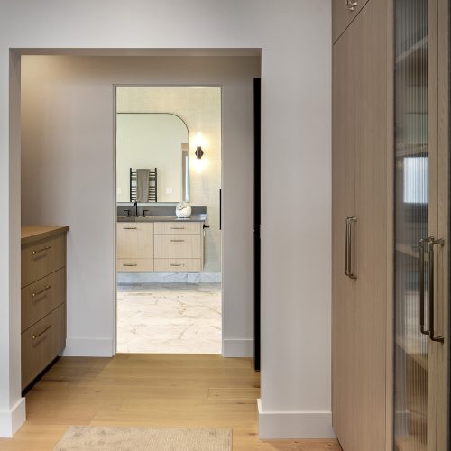 A modern walk-in closet with wood cabinets and drawers leads to a bathroom with a vanity, marble flooring, and a round mirror above the sink. The space is well-lit with natural and artificial light.