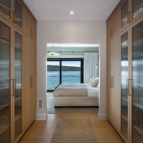 A modern walk-in closet with wooden cabinets leads to a bedroom featuring a large bed and a wall of windows overlooking a body of water and trees outside.