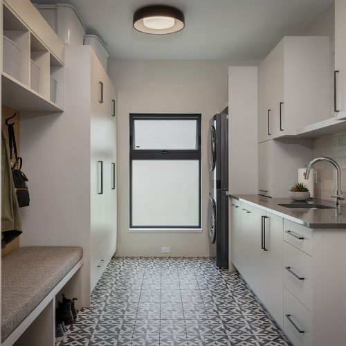 Modern laundry and mudroom with patterned tile floor, built-in bench, hooks, cabinets, stacked washer and dryer, gray countertops, white tile backsplash, sink, and a frosted window letting in natural light.