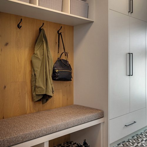 A mudroom with geometric floor tiles, a cushioned bench, and open cubbies with baskets above. A green jacket and black bag hang on hooks. Shoes and slippers are neatly placed below the bench.